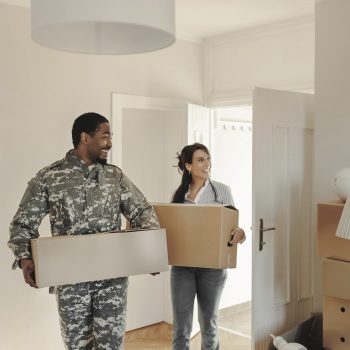 Happy wife and her african american army veteran husband in camouflage clothing with cardboard boxes in new house at moving day. First time home buyers.